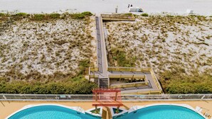 An aerial view of a beach with two swimming pools, a wooden walkway, and a lifeguard tower.