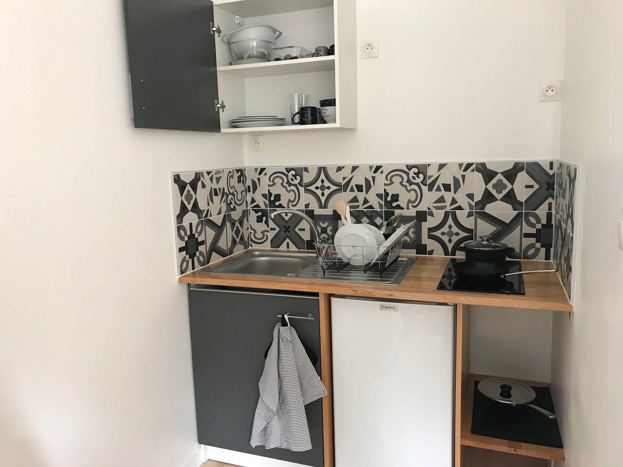 A compact kitchen with a sink, a stove, and a small refrigerator. There is a dish rack above the sink and a towel hanging on the refrigerator door.