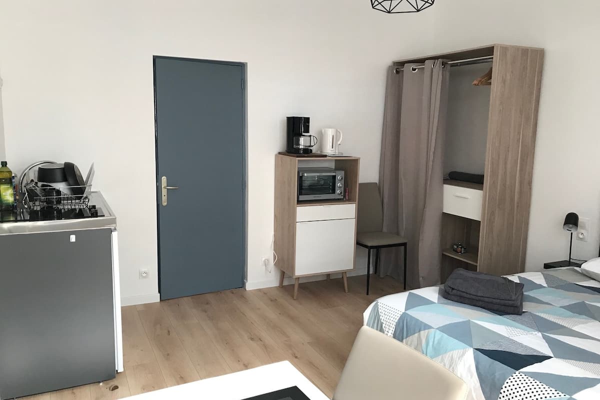 A compact hotel room with a kitchenette, a bed with a geometric patterned duvet, a chair, a desk with a microwave, and a wardrobe with a curtain.