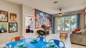 Un coin repas avec une table et des chaises bleues, un canapé avec des coussins et une grande fenêtre donnant sur l’extérieur.
