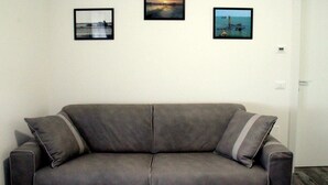 Un sofá gris con dos almohadones, una pared blanca con tres cuadros enmarcados y una puerta cerrada.