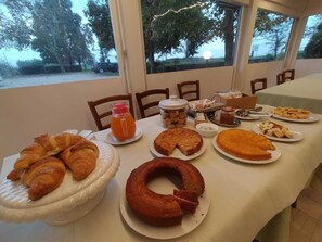 Een ontbijtbuffet met gebak, een fles sinaasappelsap en diverse desserts op een tafel bij een raam met uitzicht op bomen.