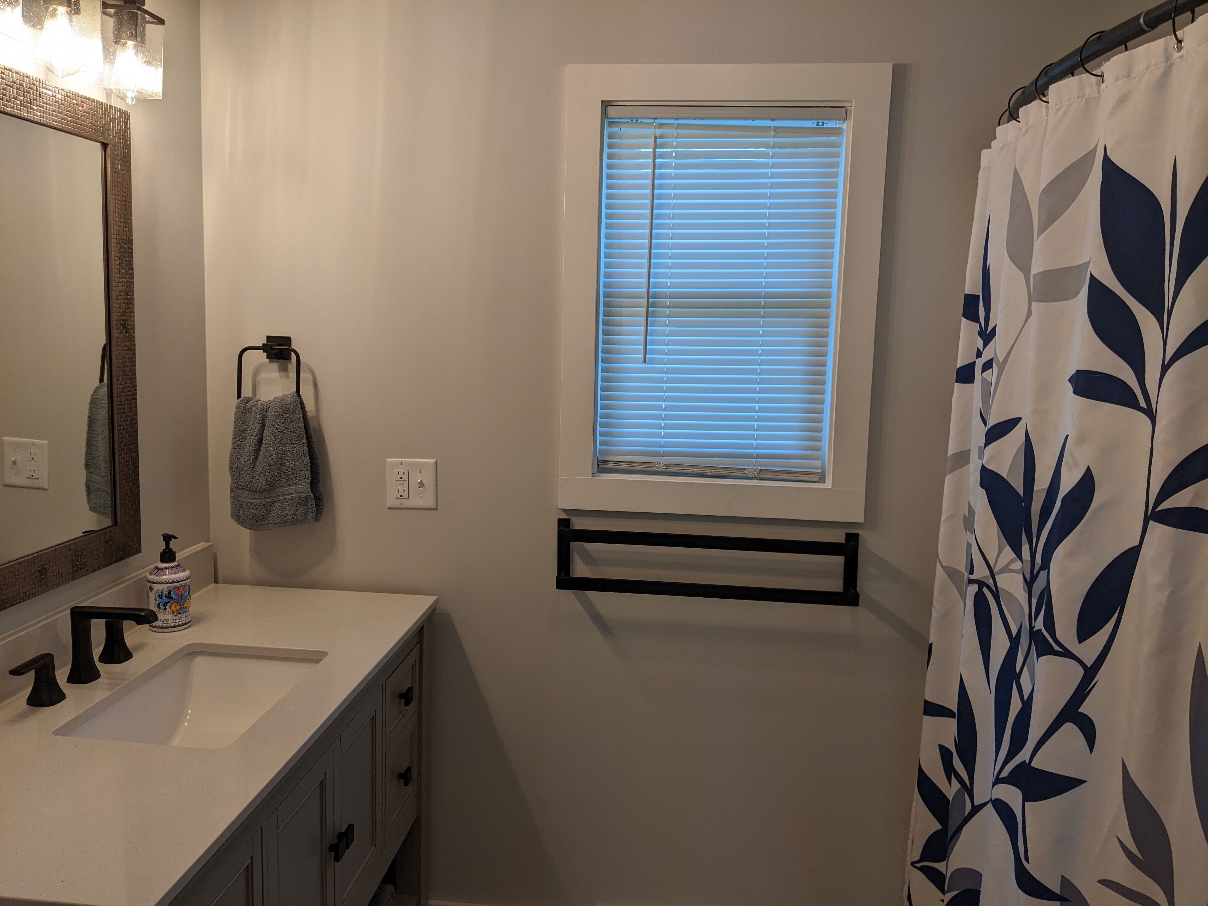 Un bagno con un lavandino bianco, uno specchio con cornice decorativa, un porta asciugamani con un asciugamano, una finestra con persiane e una tenda da doccia con motivo a foglia.