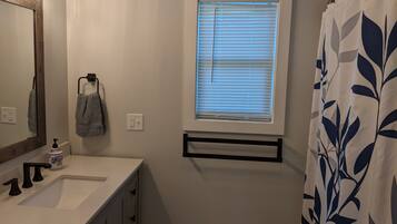 Un bagno con un lavandino bianco, uno specchio con cornice decorativa, un porta asciugamani con un asciugamano, una finestra con persiane e una tenda da doccia con motivo a foglia.