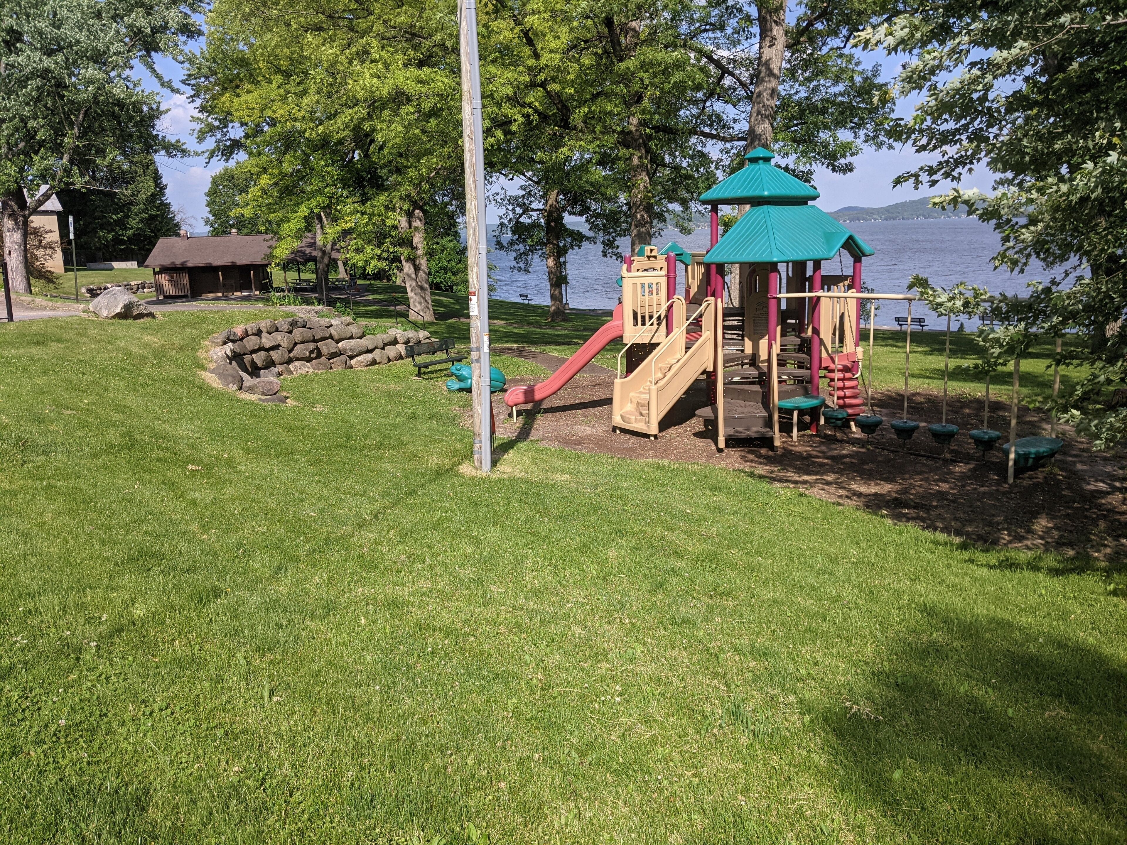 Ein Spielplatz mit Rutsche und Kletterbereich, umgeben von Gras und Bäumen, in der Nähe von Wasser.