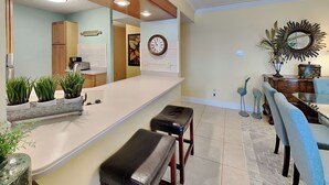 A modern kitchen with a white countertop, wooden cabinets, and a built-in oven. There is a dining area with a glass table and chairs, and a decorative mirror on the wall. - Fort Walton Beach Condo with Beachfront Balcony, Views of The Gulf of Mexico, an (Fort Walton Beach)