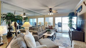 A spacious living room with a large sofa, a coffee table, and a TV. There are two lamps, a ceiling fan, and a balcony with a chair. - Fort Walton Beach Condo with Beachfront Balcony, Views of The Gulf of Mexico, an (Fort Walton Beach)