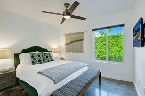 A bedroom with a bed, a ceiling fan, a nightstand, a lamp, a window with a view of greenery, and a wall-mounted TV. - Casa Azul - Mid-Century Oasis w/ Private Pool & Mountain Views (Palm Springs)