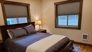 Un dormitorio con una cama grande, cabecera de madera, dos mesitas de noche con lámparas y ventanas con persianas.