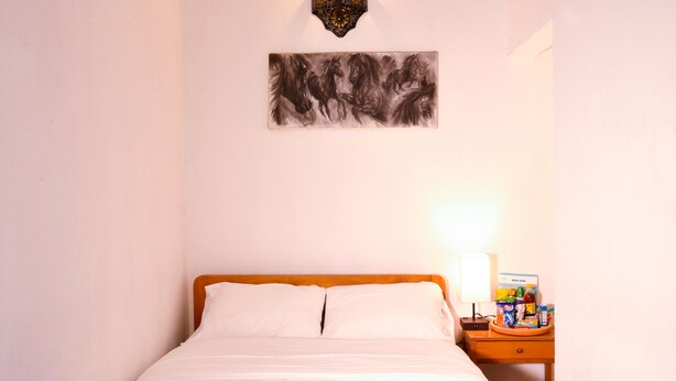 Un lit bien fait, avec une literie blanche et bleue, une tête de lit en bois et une table de chevet sur laquelle se trouvent une lampe et un plateau avec divers objets.