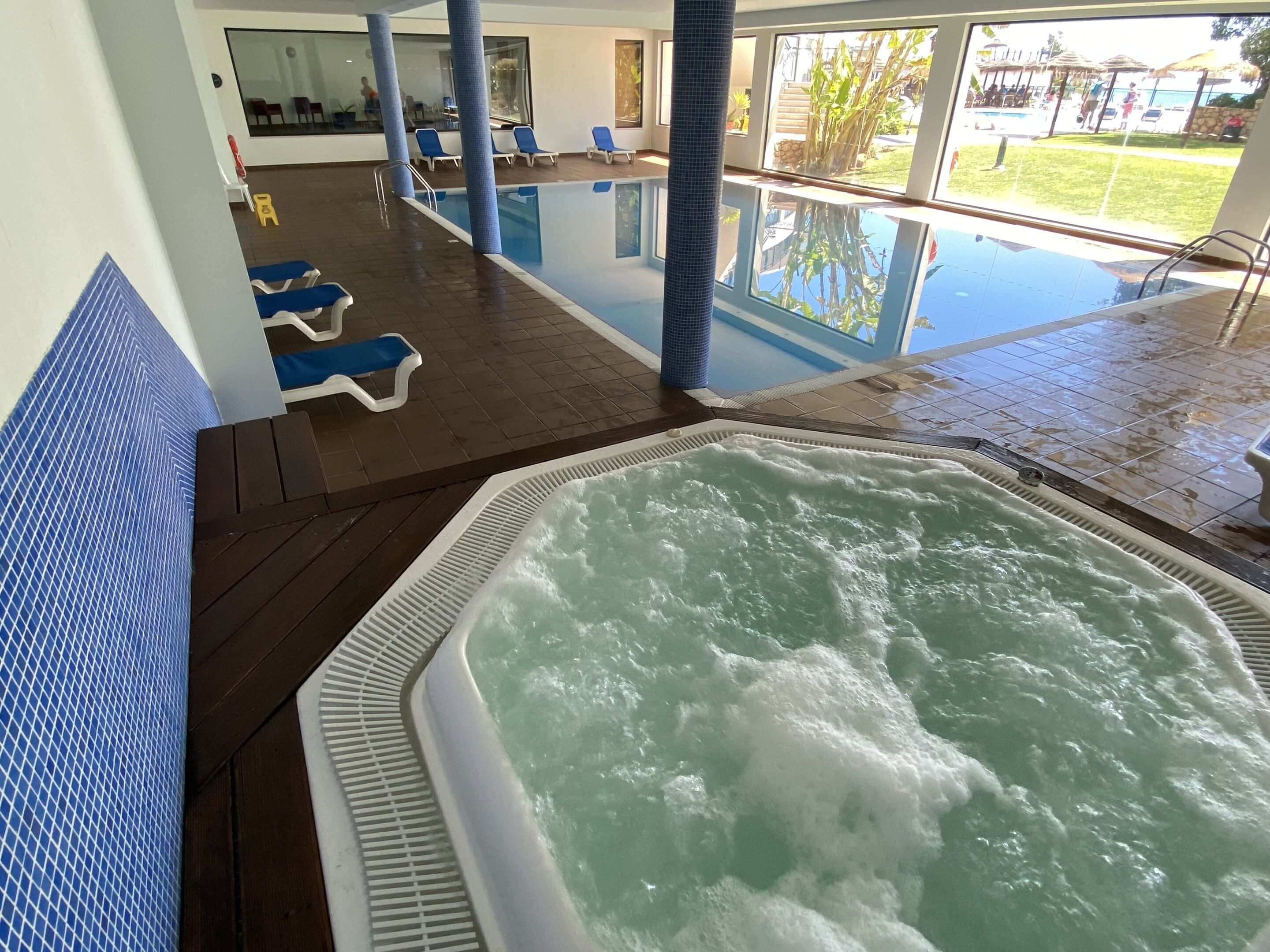An indoor swimming pool with a hot tub, lounge chairs, and a view of the outdoor pool area.