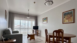 Uma sala de estar com sofá, uma mesa de jantar com cadeiras, televisão em um móvel e uma janela grande com cortinas.