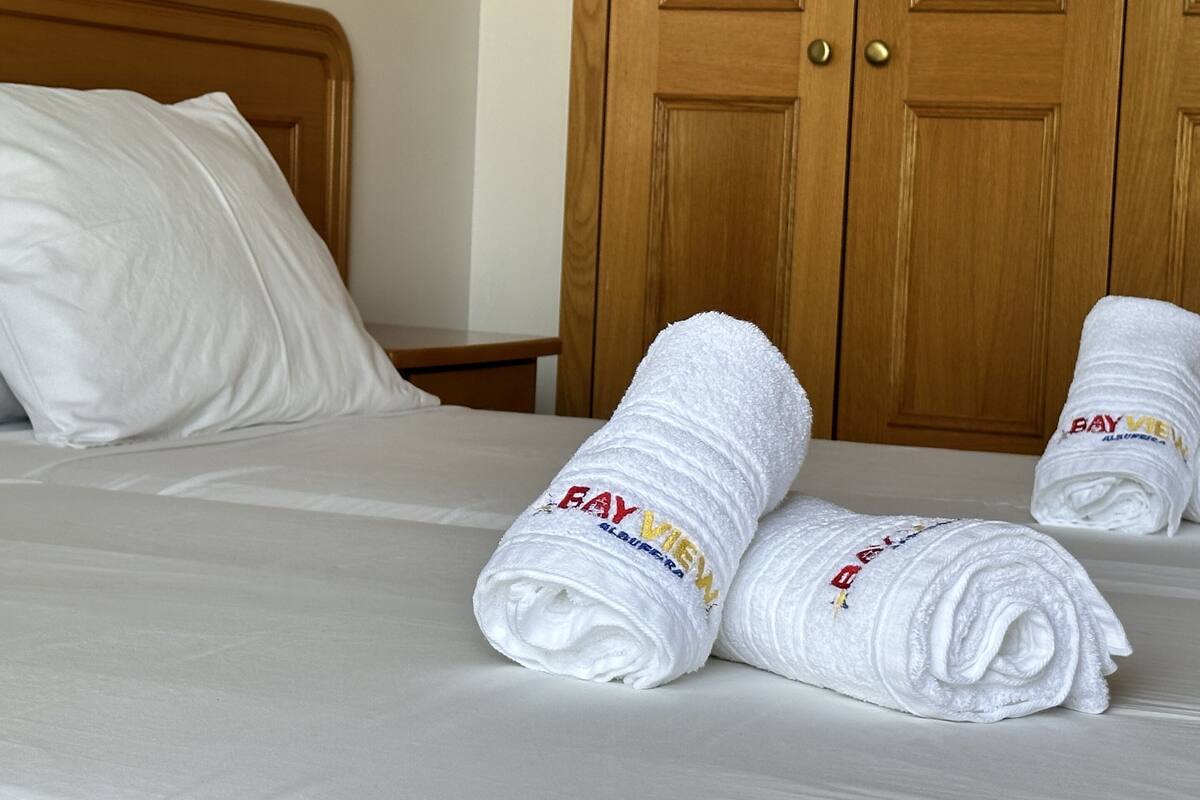 A neatly made bed with white linens and two rolled towels with embroidered text.
