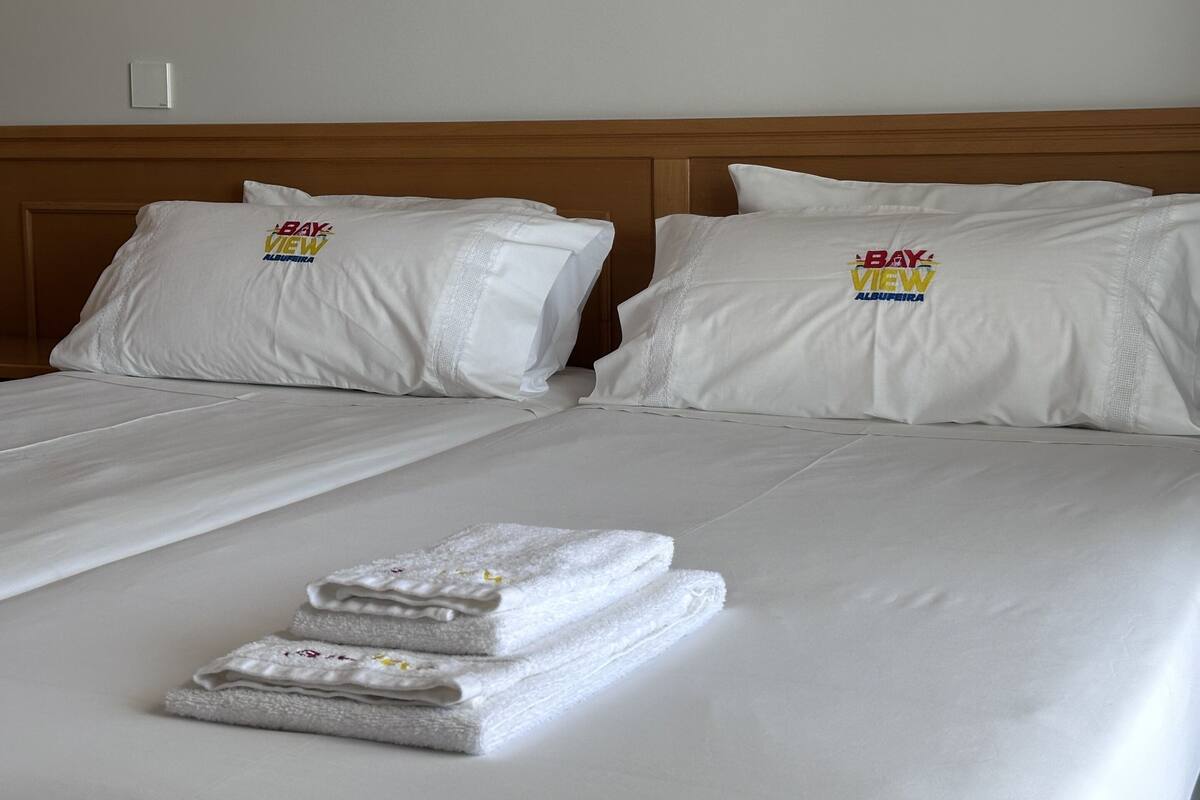 A hotel room with two beds, each with white bedding and pillows featuring the 'Bay View Albuquerque' logo. There are folded towels on the beds.