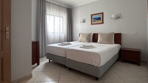 Uma cama de casal com duas almofadas, um criado-mudo, uma luminária fixada na parede e um quadro emoldurado.