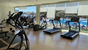 Academia com esteiras, elípticos e bancos de peso. Grandes janelas oferecem vista para uma piscina e área de estar ao ar livre.