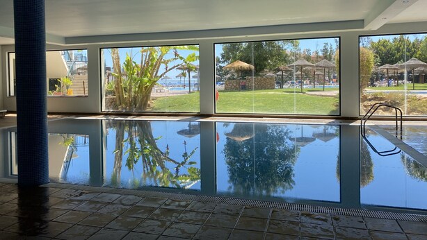 Uma grande piscina interior com vista desobstruída para área tropical externa, incluindo um espaço de descanso à beira da piscina e um gramado.