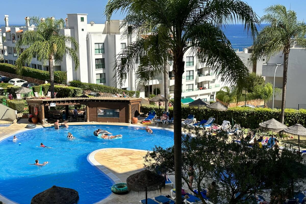 A swimming pool surrounded by lounge chairs and umbrellas, with palm trees and a building in the background.