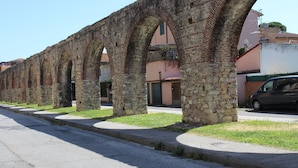 A street lined with stone arches, a car parked on the side, and buildings in the background. - Camera MINERVA (Pisa)