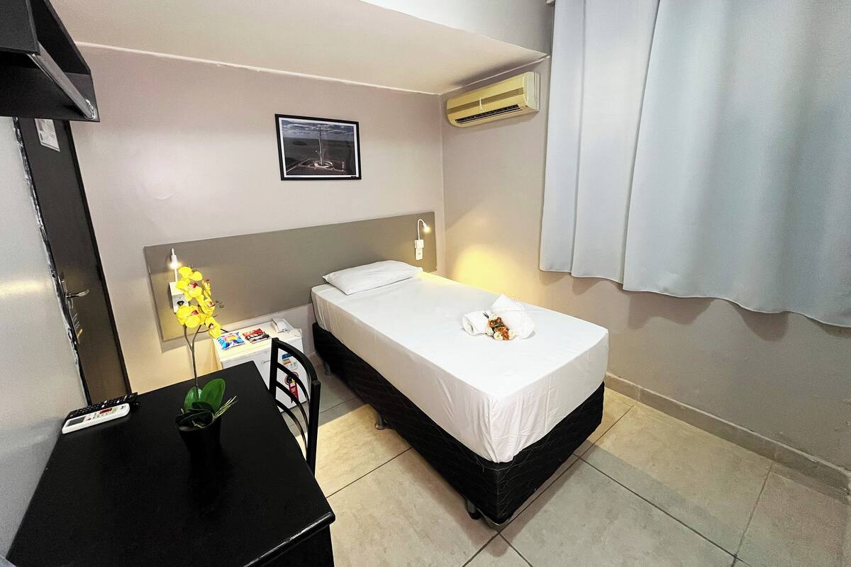 Ein Hotelzimmer mit einem Einzelbett, einem Schreibtisch mit Telefon, einem kleinen Tisch mit Pflanze und einer Klimaanlage.