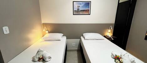 A hotel room with two single beds, each with a pillow and a towel. There is a framed picture on the wall above the beds.