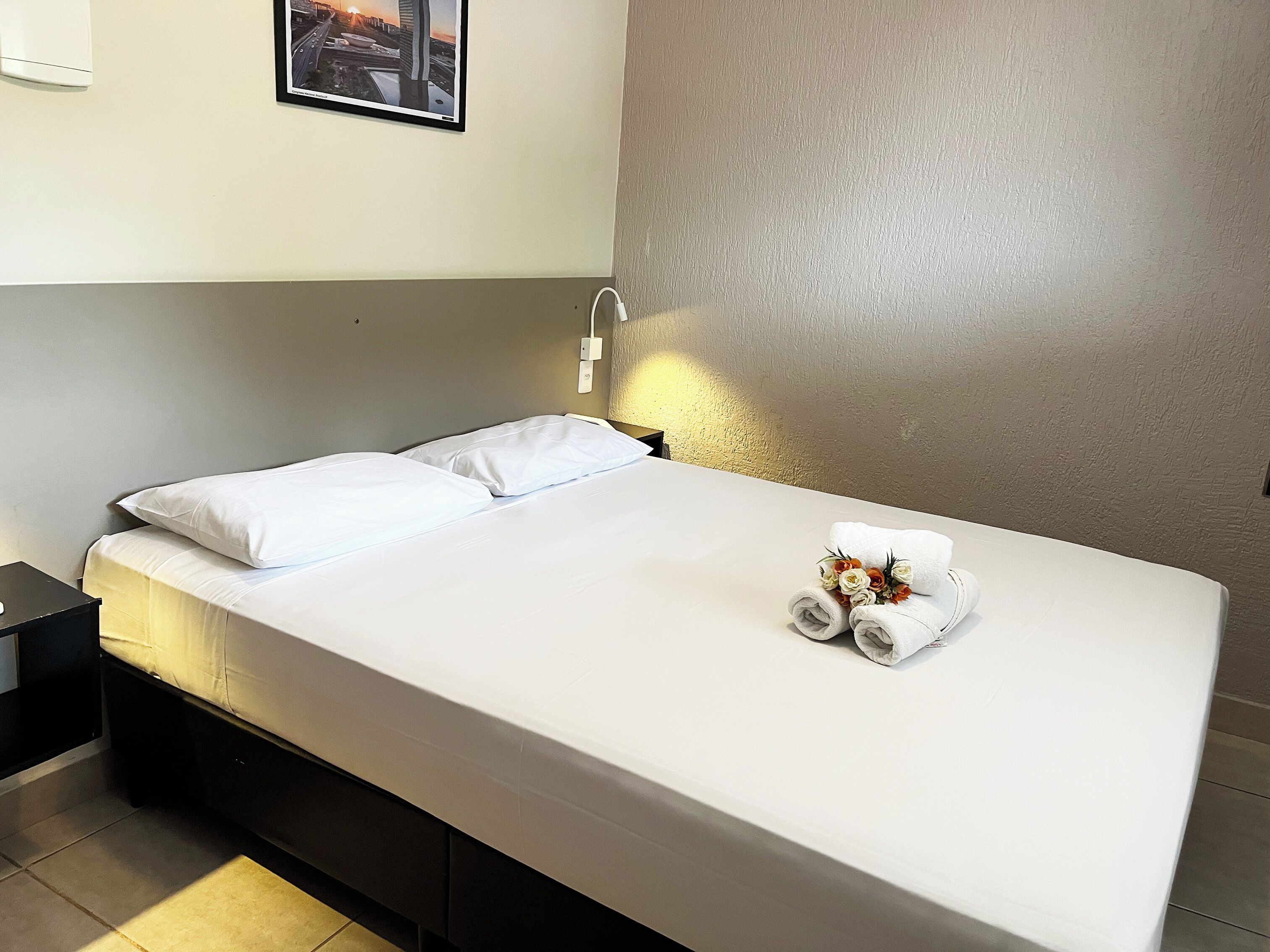 Una cama bien hecha con sábanas blancas y una toalla con flores, una mesita de noche y una lámpara de pared.