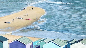 Una playa con orilla arenosa, algunas personas y algunos barcos. Se observan edificios con techos verdes al frente.