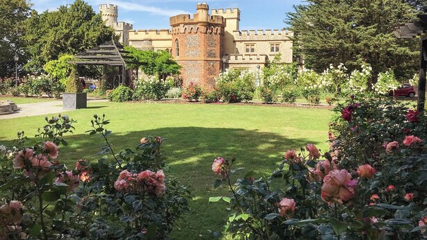 Un edificio al estilo de castillo con una torre central, rodeado de un jardín bien cuidado con rosales.