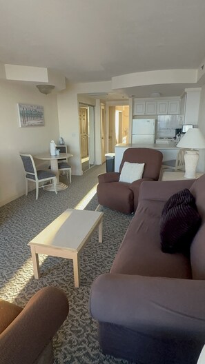 Een woonkamer met een bruine bank, een witte salontafel en een kamerbreed tapijt. Er is een eethoek met een tafel en stoelen, en een keuken op de achtergrond.