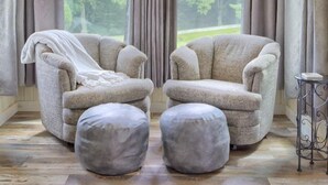 Ein gemütliches Zimmer mit zwei Sesseln, Poufs, einem kleinen Tisch und einem großen Fenster mit Vorhängen.