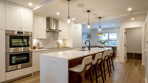 A modern kitchen with stainless steel appliances, a large island with bar stools, pendant lighting, and a view to the dining area.