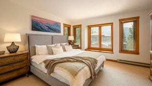 A bedroom with a large bed, wooden nightstands, a painting above the bed, and three windows with blinds.