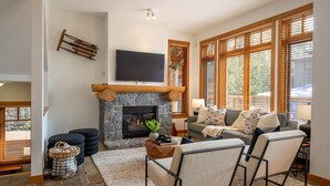 A living room with a stone fireplace, a flat-screen TV mounted above it, a sofa with cushions, and large windows with wooden blinds.