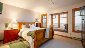 Un dormitorio con cama de madera, mesitas de noche, un banco y ventanas grandes con persianas.