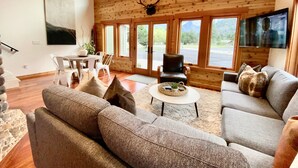 Ein geräumiges Wohnzimmer mit einer großen Couch, einem Couchtisch und einem Fernseher. Es gibt eine Holzbalkendecke mit großen Fenstern und einer Dekoration mit Hirschgeweih.