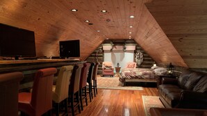 A cozy attic room with a wooden ceiling, a bar area with stools, a bed, and a leather couch.