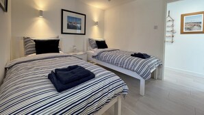A hotel room with two beds, each with striped bedding, a bedside table, and a framed picture on the wall. - A beautiful coastal cottage in characterful Portmahomack. (Portmahomack, near Dornoch)