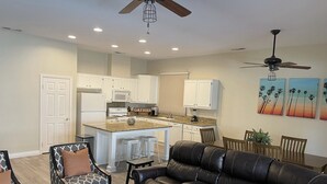 A modern kitchen with white cabinets, a central island, and a dining area with a black leather couch and patterned chairs. - Desert Oasis salt water Pool/spaHome, Gated Community w/LAKE (Indio)