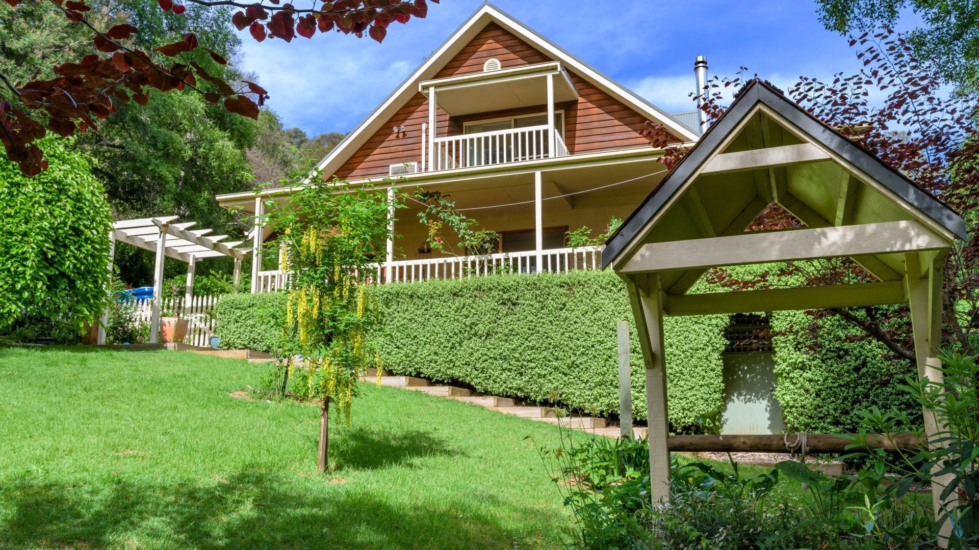 A two-story wooden house with a balcony, surrounded by a well-maintained garden and a pergola.