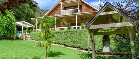 A two-story wooden house with a balcony, surrounded by a well-maintained garden and a pergola.