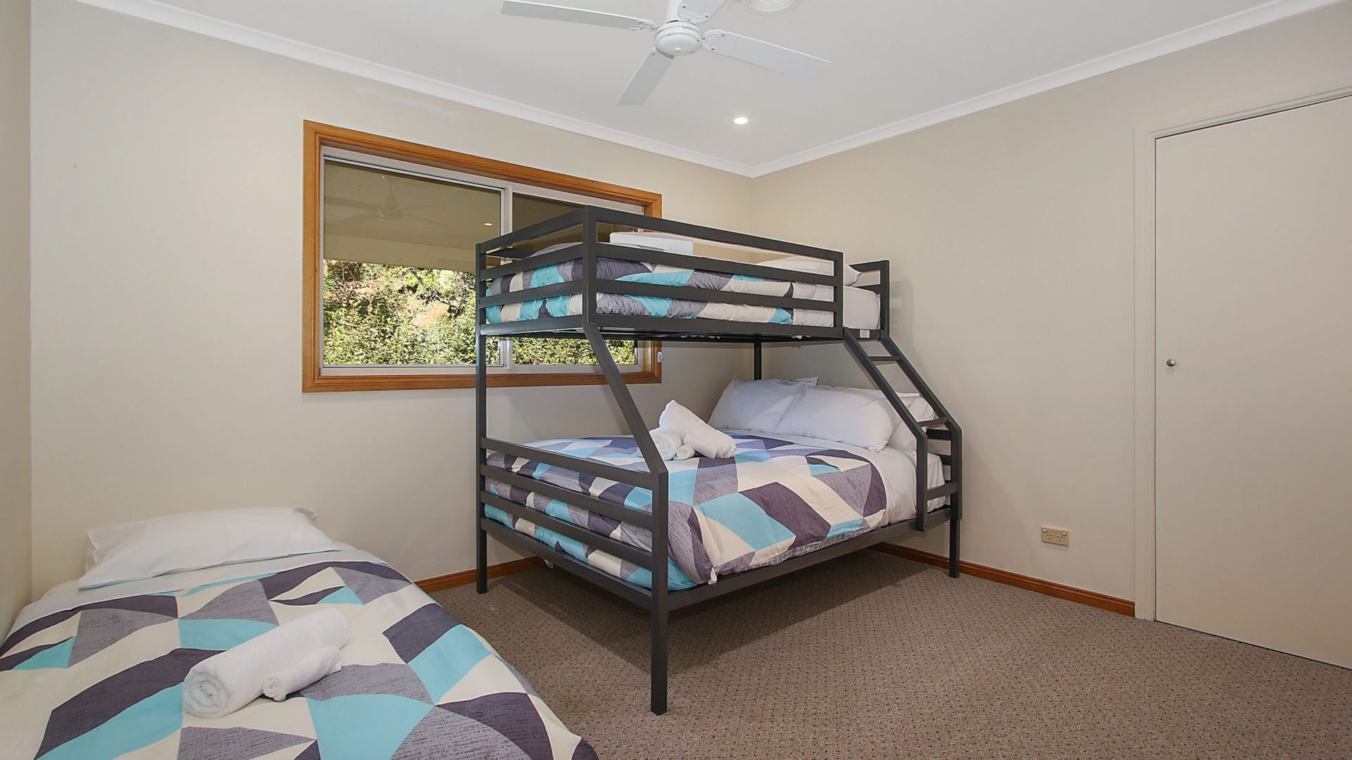A room with a bunk bed, a single bed, a window with a view of greenery, and a ceiling fan.