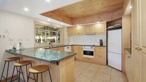 A modern kitchen with wooden cabinets, a granite countertop, a built-in oven, and a refrigerator. There are four bar stools and a window providing natural light. - Greenlea (BRIGHT)