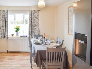 A dining room with a table set for four, a radiator, a window with curtains, and a refrigerator. - Stepping Stone Cottage (Bacton, near North Walsham)