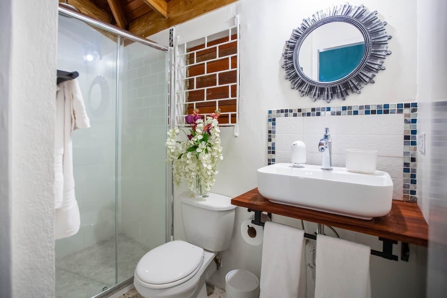 A bathroom with a glass shower enclosure, a toilet, a sink with a mirror above it, and a vase with flowers on the wall.