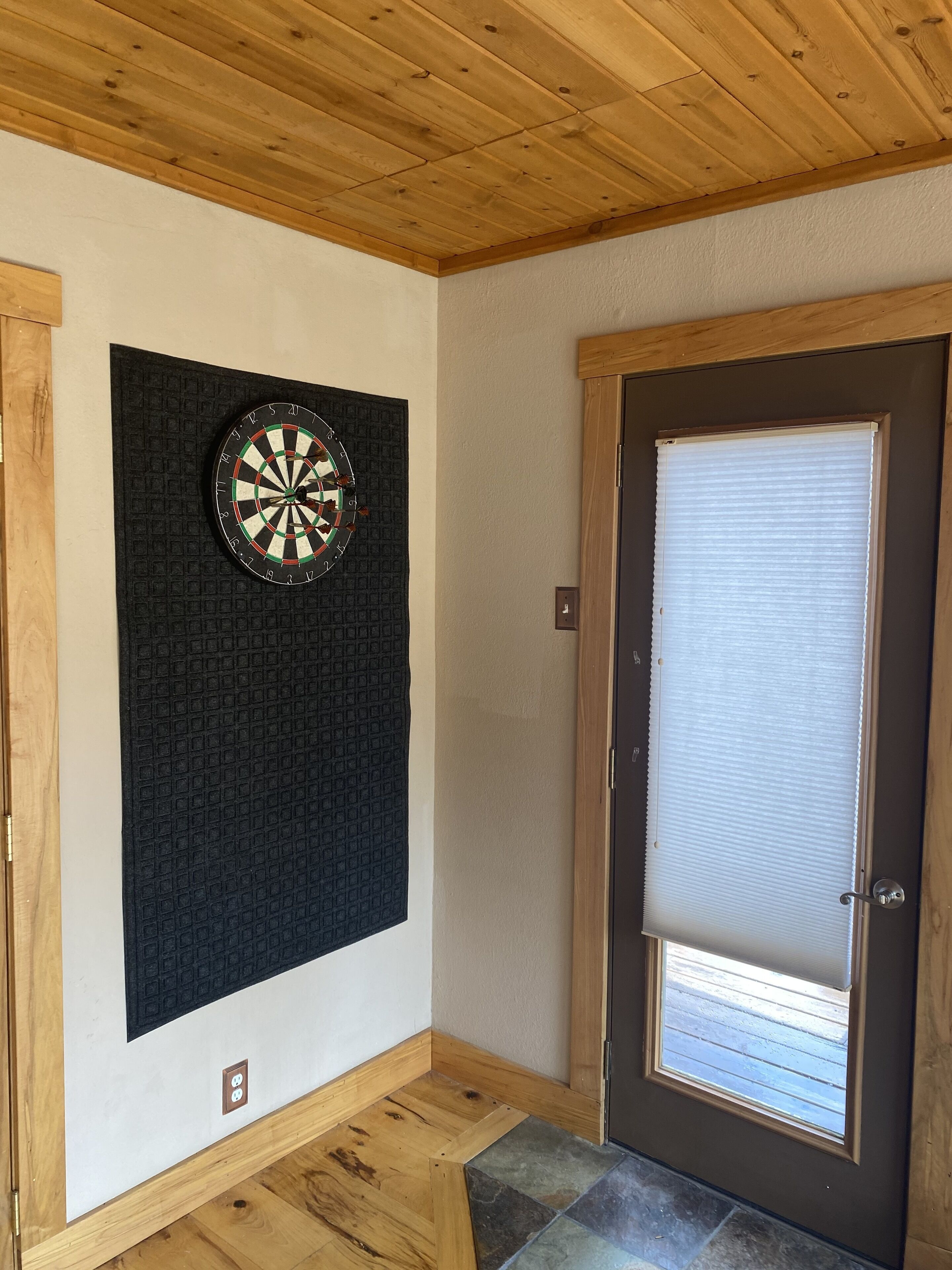 A room with a dartboard mounted on a textured wall, a wooden door with a window, and a tiled floor.