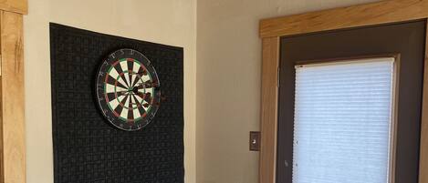A room with a dartboard mounted on a textured wall, a wooden door with a window, and a tiled floor.