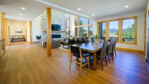 A spacious living area with a wooden dining table, a stone fireplace, and large windows offering a view of the outdoors. - The Ledges | Ski Getaway | Hot Tub & Theater Room (Eden)