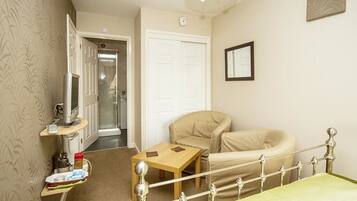 A hotel room with a bed, a chair, a small table, a shelf with a TV, and a door leading to another room.