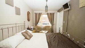 A hotel room with a bed, a desk, a chair, a small table, a window with curtains, a wall clock, and a television mounted on the wall. - Professional Bed & Breakfast en-suite double rooms £80 per night per room. (Warwick)
