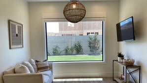 A modern living room with a large window, a white sofa, a wicker pendant light, a flat-screen TV, and a small side table with a plant.